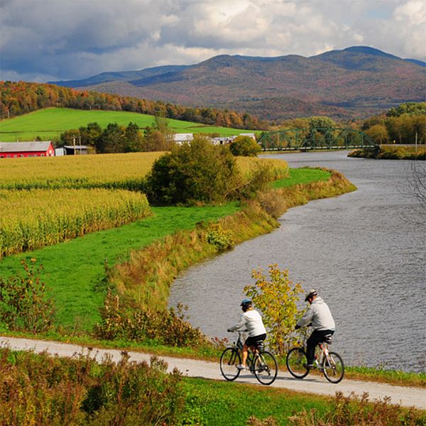 Photo of bicyclists on a rec path surrounded by a fall landscape with the Missiquoi River and mountains.